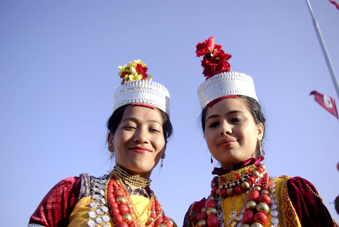 Two women from Meghalaya, India, wearing traditional attire with reds and yellows, jewelry resembling beads around their necks, and a white hat with red flowers, smiling and looking down.