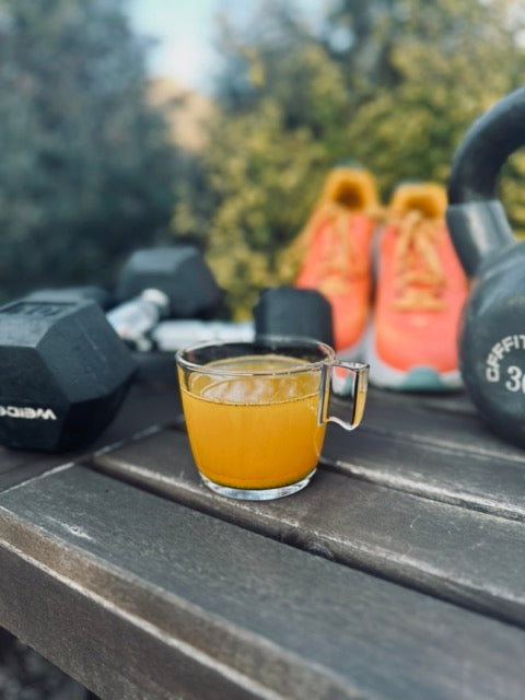 Simple Origins Superfoods Immunity Boosting Medley is shown in a glass cup on a rustic bench with dumbells, kettlebells and sneakers around it. Light and dark green bushes and trees are in the background. Nourish and Move.  