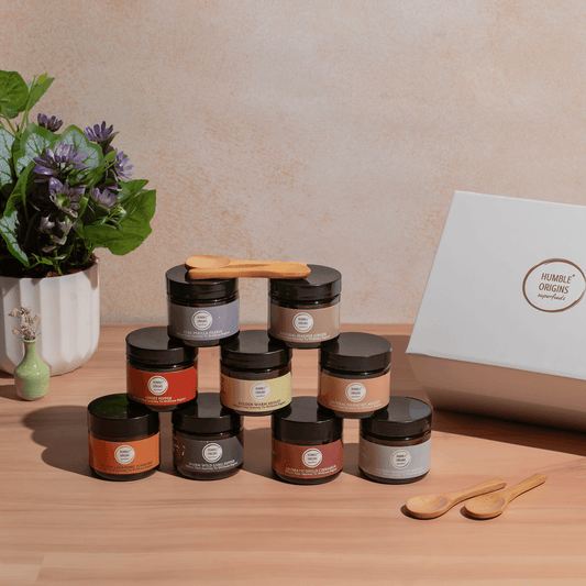 Assorted jars with lids on a wooden surface, with a plant and box in the background.