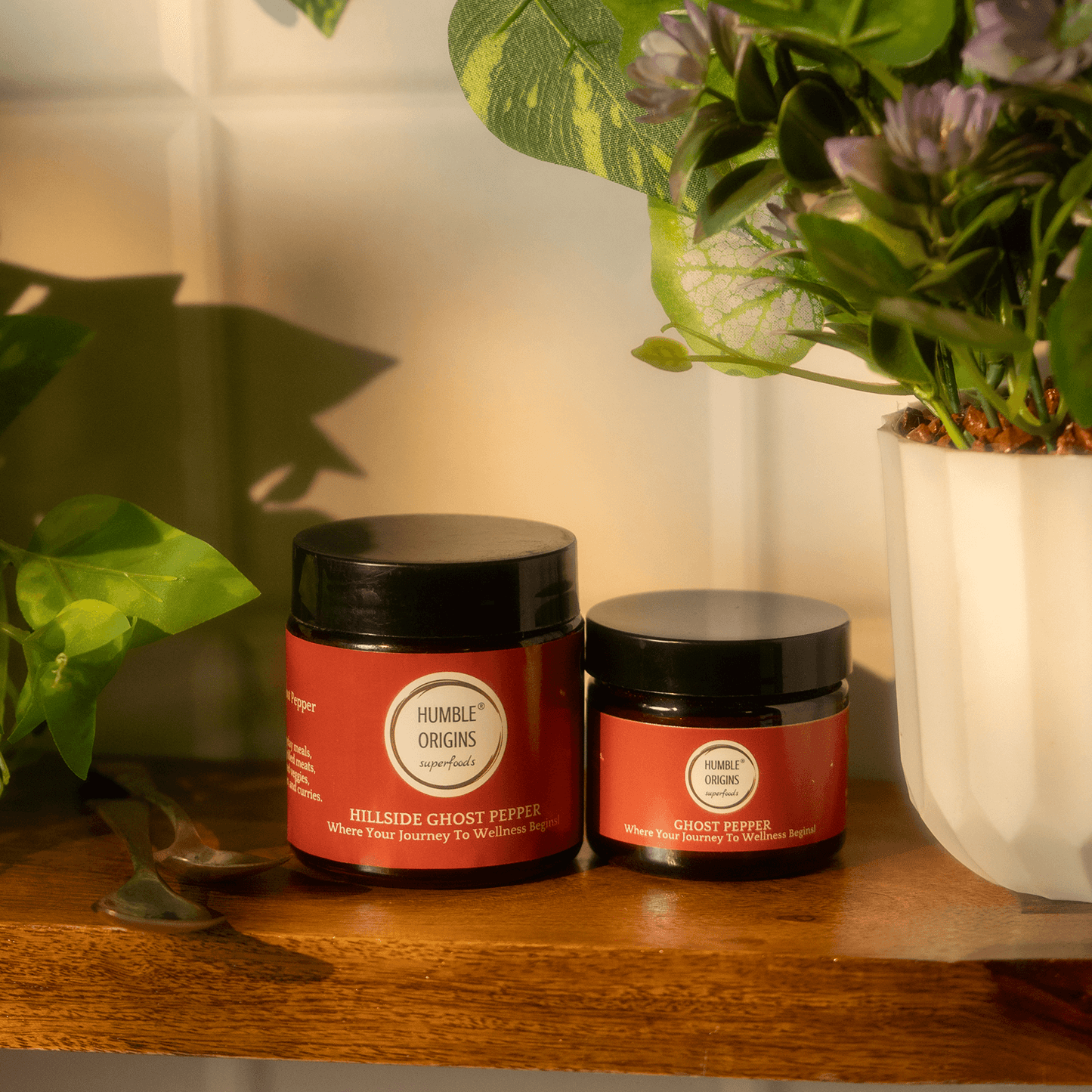 Two jars of 'Humble Origins' on a wooden surface with plants in the background